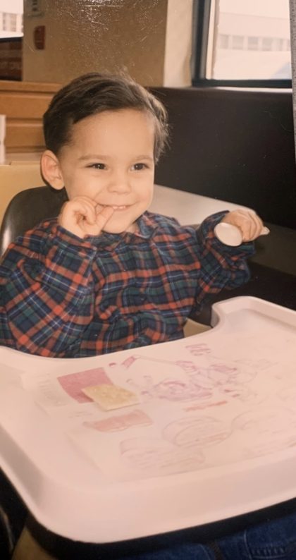 Andrew as a toddler in a high chair, grinning, with a cracker on the tray